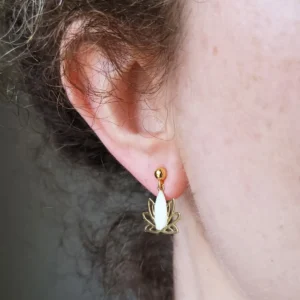 Clous d'oreilles dorés avec un pendant en forme de lotus ajouré de couleur doré et blanc, portés.