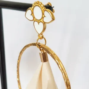 Boucles d'oreilles pendantes avec des supports en formes de 3 coeurs colorés en blanc et dorés. Ces supports ont un anneau martelé et un pompon en imitation satin de couleur beige à l'intérieur.