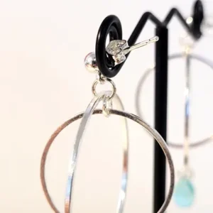 Support des boucles d'oreilles argentées Galilée composées de 2 cercles imbriqués de tailles différentes et d'une goutte en verre translucide colorée. Le support est un clou d'oreille en argent.