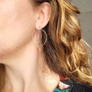 Boucles d'oreilles argentées avec 2 cercles imbriqués de tailles différentes et une perle de verre de Bohème en forme de goutte de couleur pêche. Le support est un clou d'oreille en argent. Photo avec la boucle d'oreille portée.