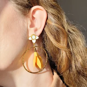 Vue portée des boucles d'oreilles pendantes avec des supports en formes de 3 coeurs colorés en blanc et dorés. Ces supports ont un grand anneau martelé et un pompon en imitation satin de couleur moutarde à l'intérieur.